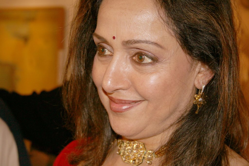 Hema Malini smiling in a red outfit and gold choker at an event in Kolkata