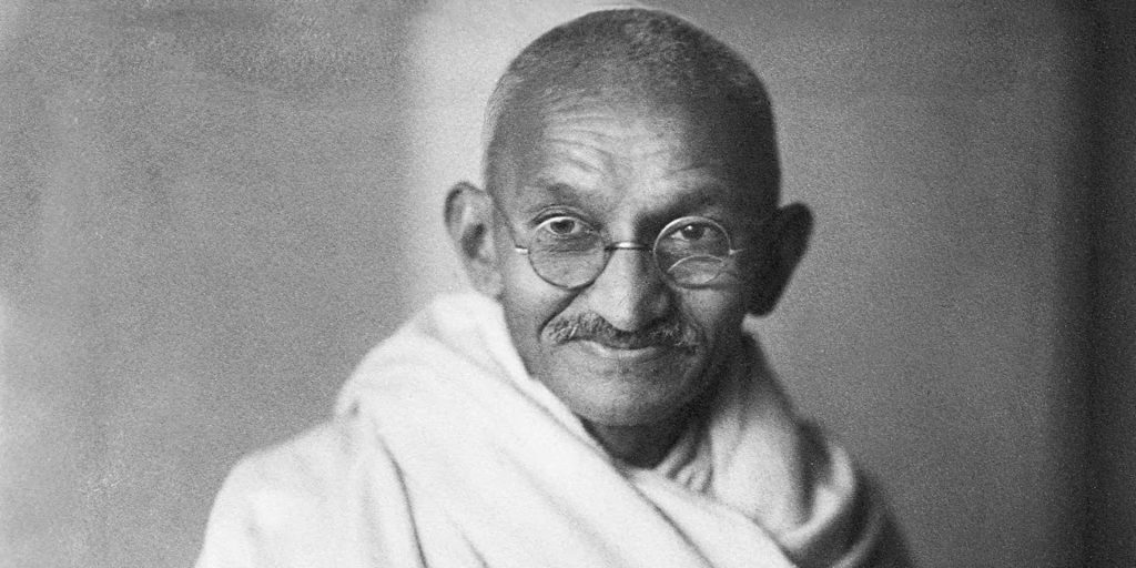 Black-and-white portrait of a smiling Mahatma Gandhi in round glasses and a white shawl