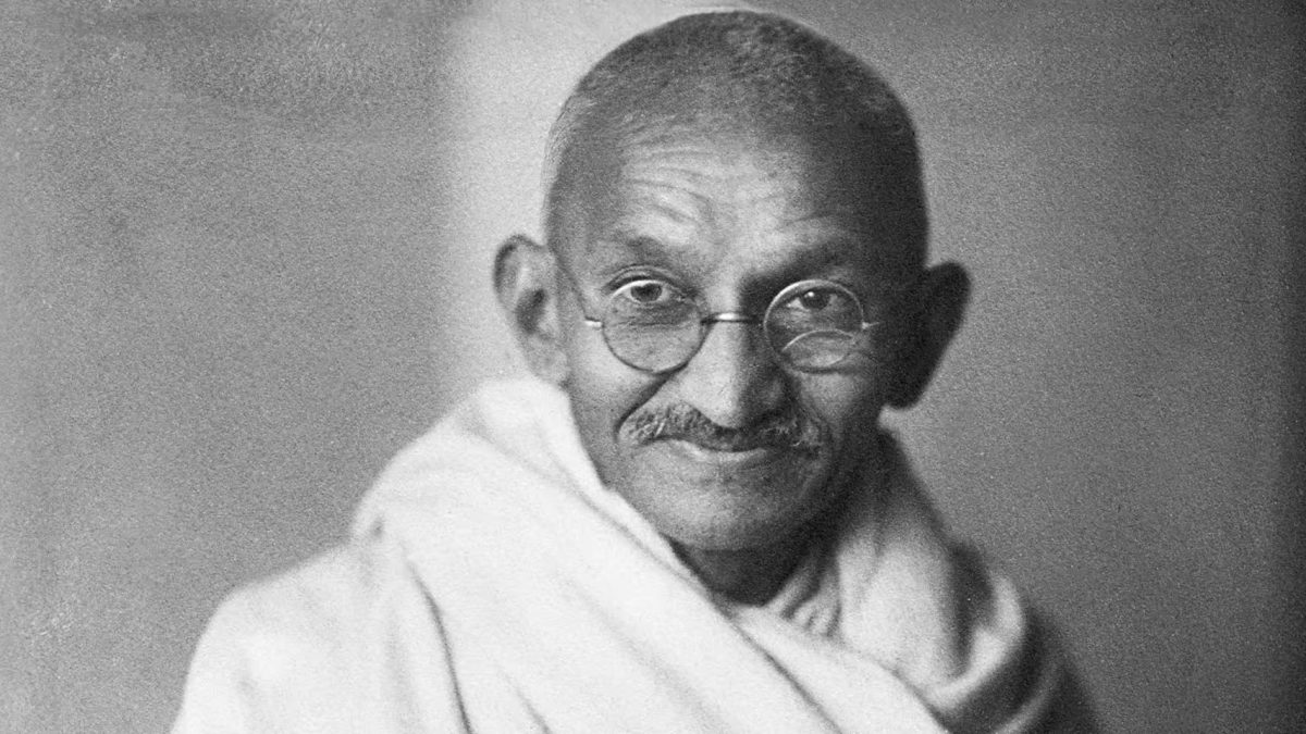Black-and-white portrait of a smiling Mahatma Gandhi in round glasses and a white shawl