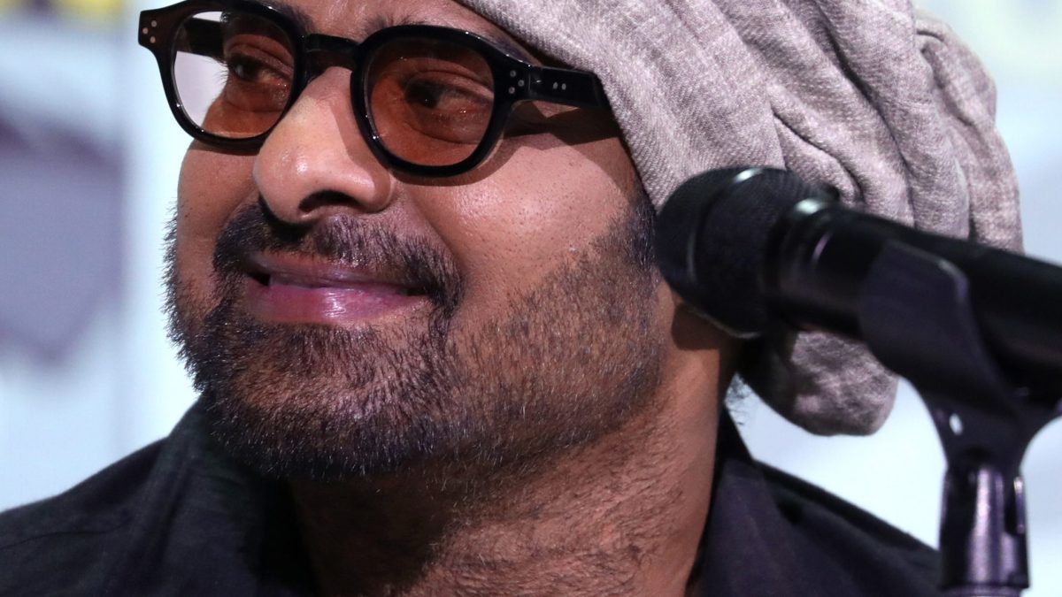 Prabhas in black-rimmed glasses and a grey head wrap speaking at a microphone at San Diego Comic-Con