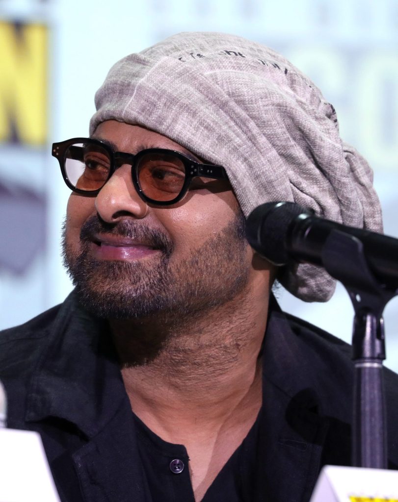 Prabhas in black-rimmed glasses and a grey head wrap speaking at a microphone at San Diego Comic-Con