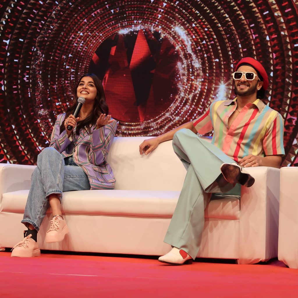Ranveer Singh in a striped shirt, red beret and sunglasses seated on stage beside a fellow guest at Vaayu 2022