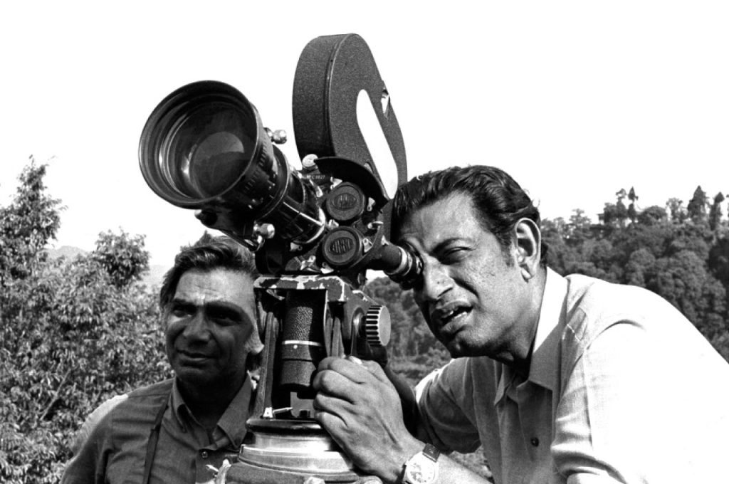 Black-and-white photo of Satyajit Ray looking through a film camera on location, with a colleague beside him