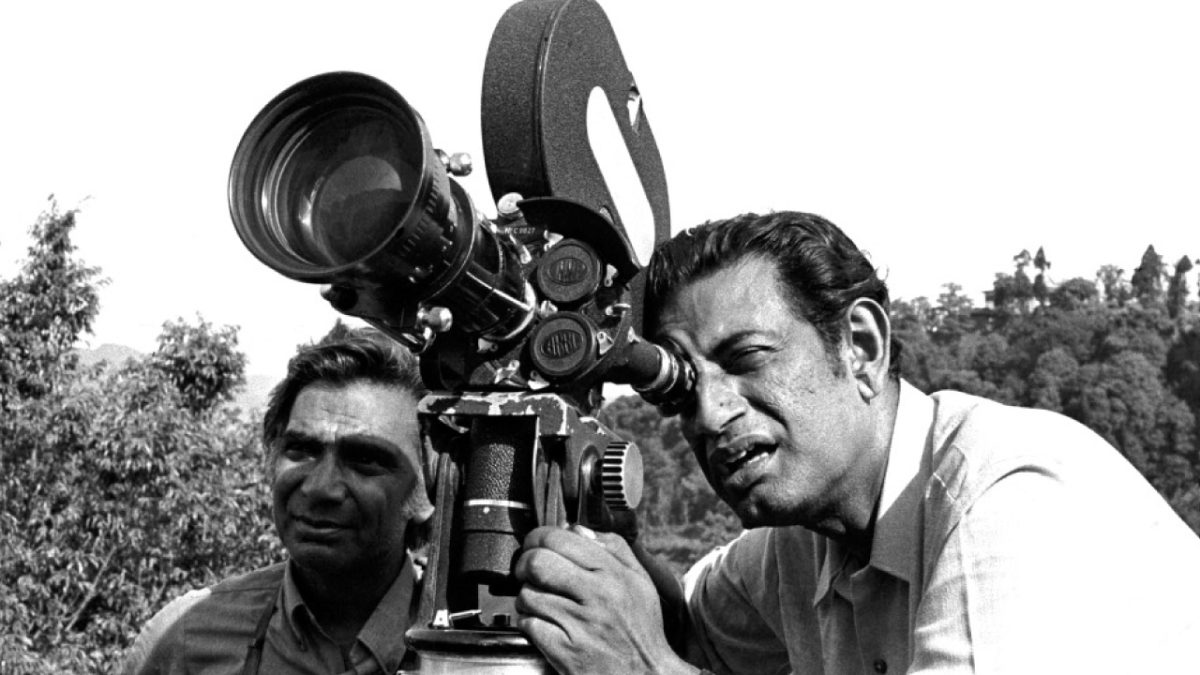 Black-and-white photo of Satyajit Ray looking through a film camera on location, with a colleague beside him