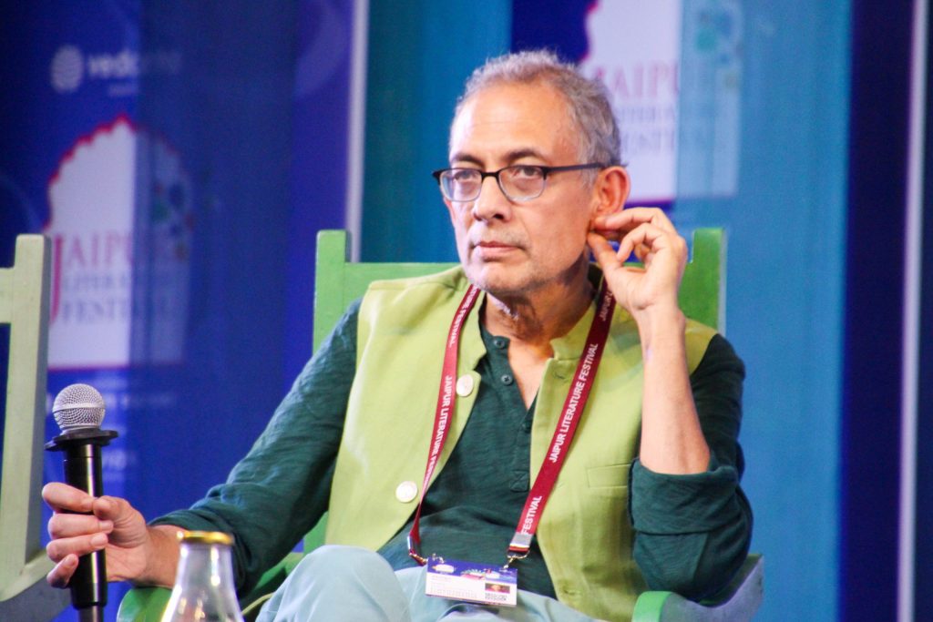 Abhijit Banerjee in glasses and a green kurta seated on stage holding a microphone