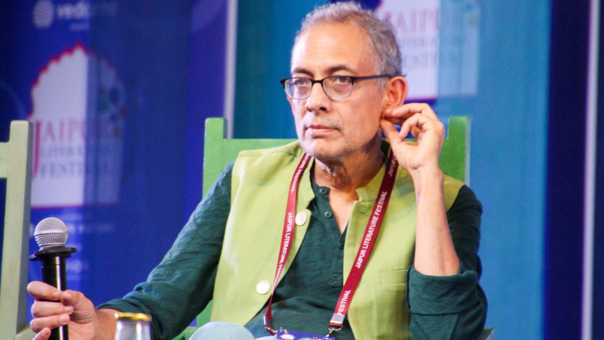 Abhijit Banerjee in glasses and a green kurta seated on stage holding a microphone