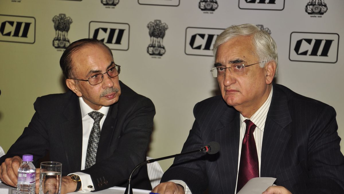 Adi Godrej in glasses and a dark suit seated beside External Affairs Minister Salman Khurshid at a conference