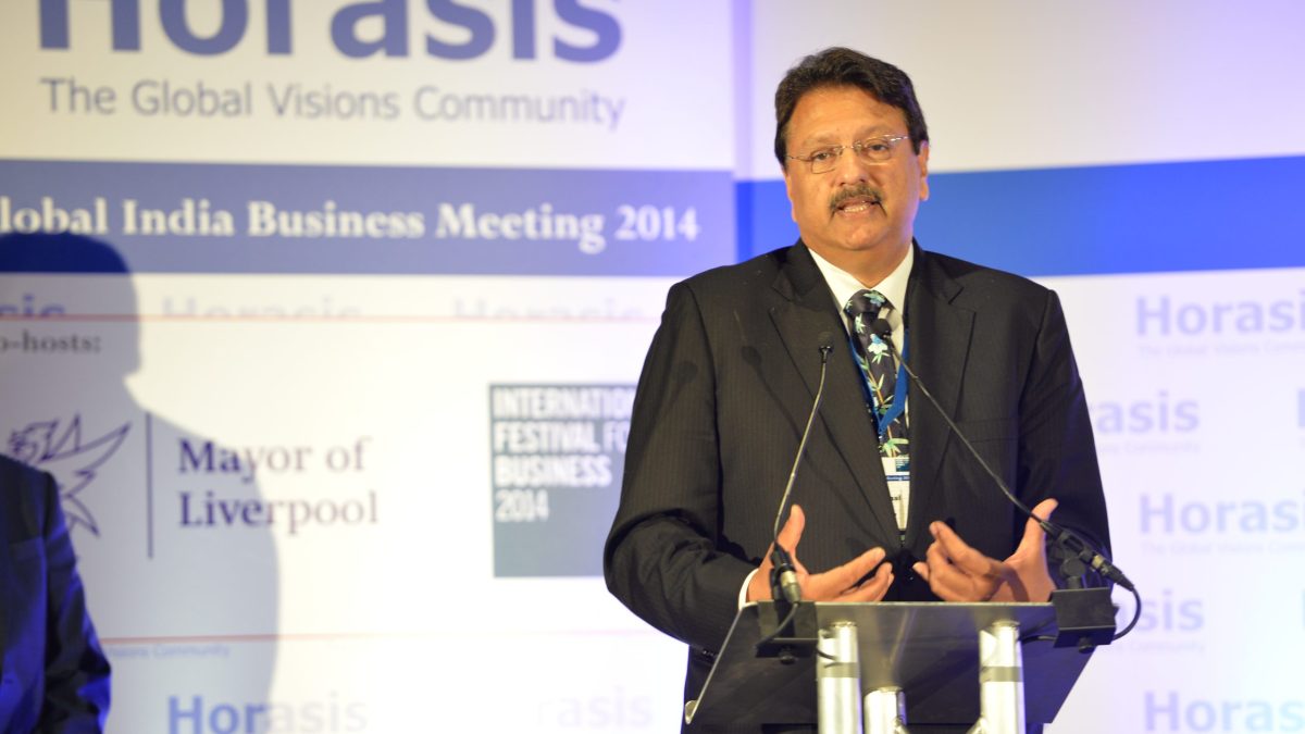 Ajay Piramal in a dark suit and red tie speaking with hands raised