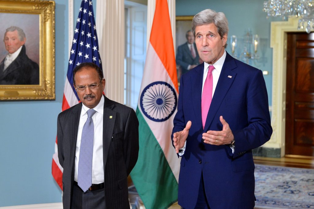 Ajit Doval in a dark suit standing beside US Secretary of State John Kerry in front of Indian and American flags