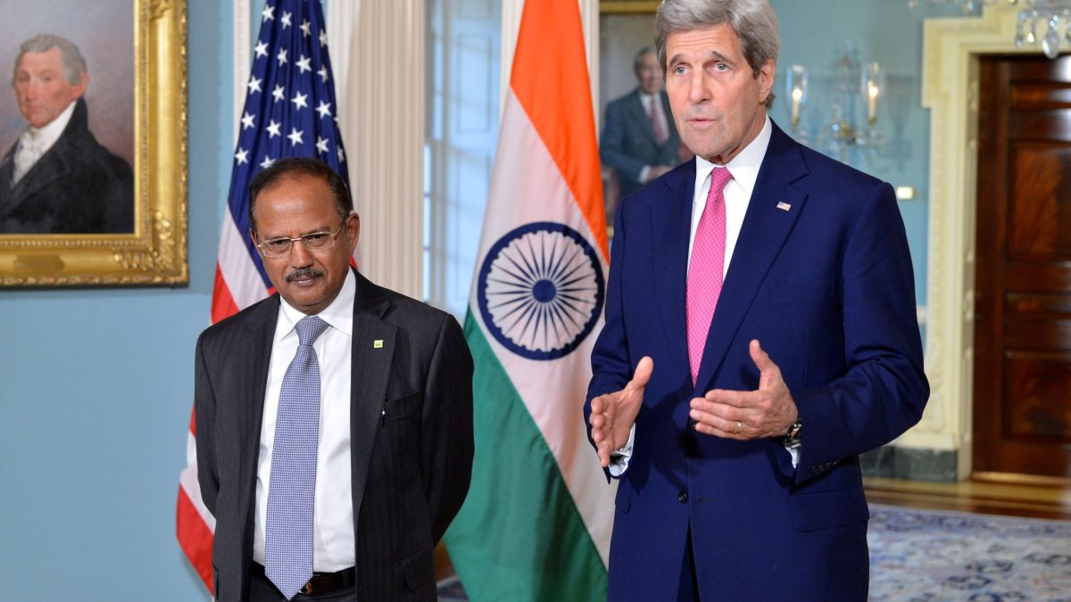 Ajit Doval in a dark suit standing beside US Secretary of State John Kerry in front of Indian and American flags