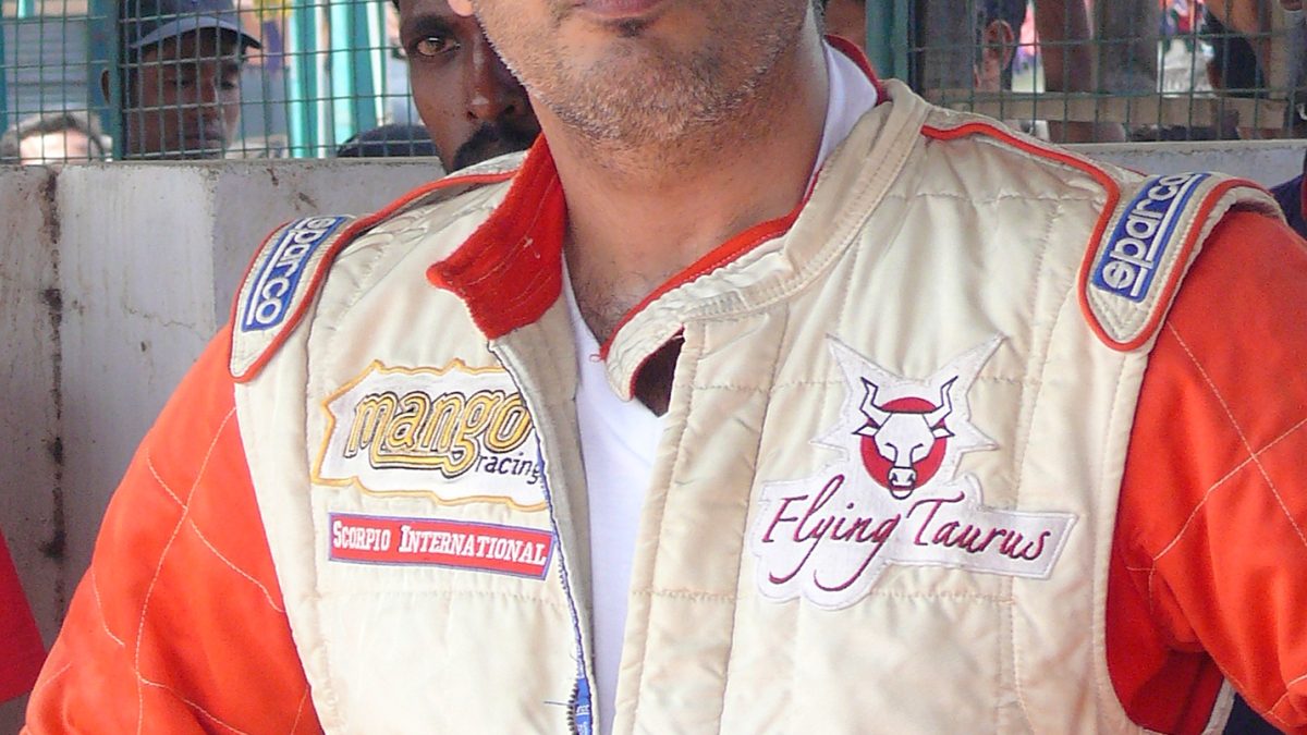 Ajith Kumar in an orange and white racing suit at a race track