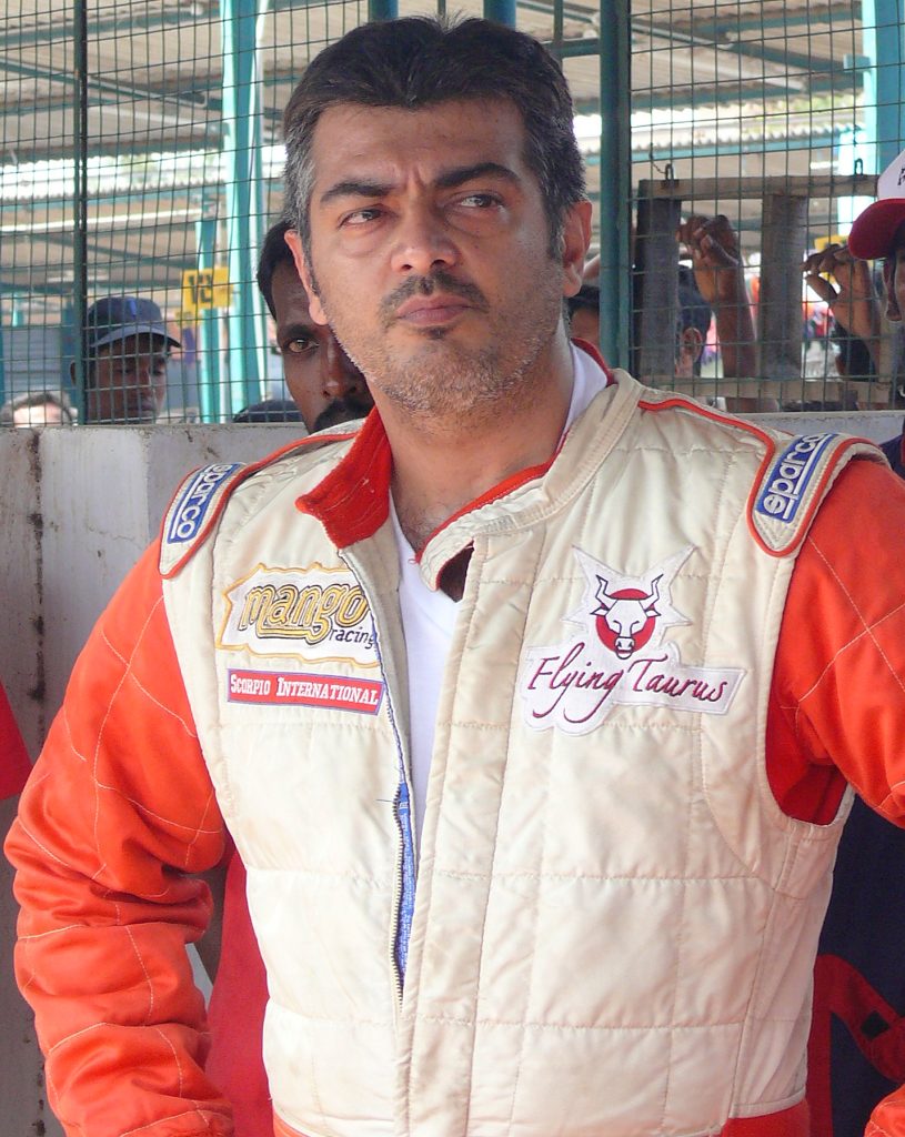 Ajith Kumar in an orange and white racing suit at a race track