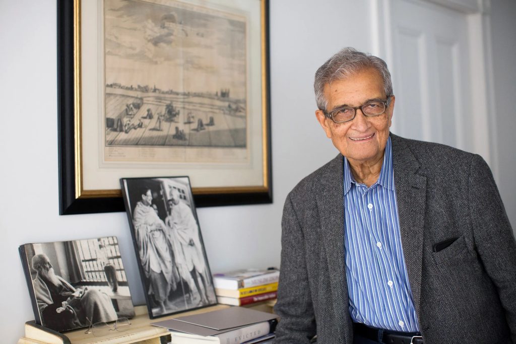 Amartya Sen smiling in a grey jacket beside framed photographs of Tagore and Gandhi