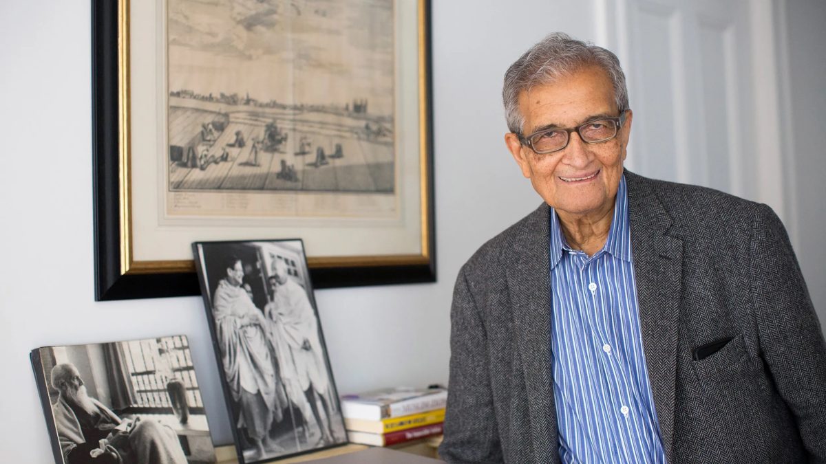Amartya Sen smiling in a grey jacket beside framed photographs of Tagore and Gandhi