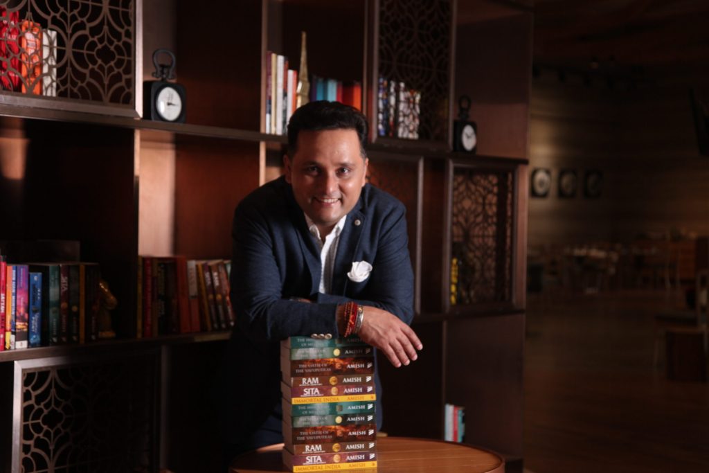 Amish Tripathi in a dark blazer leaning on a stack of his books in a library