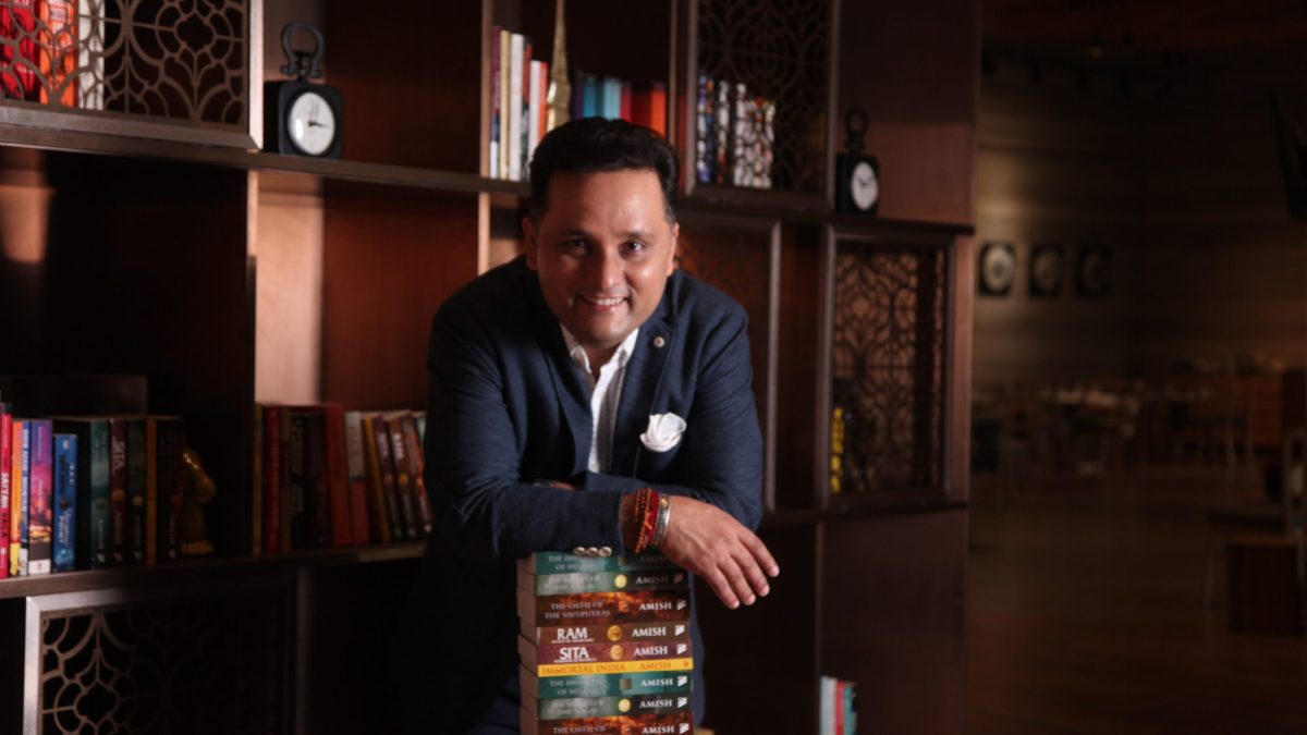 Amish Tripathi in a dark blazer leaning on a stack of his books in a library