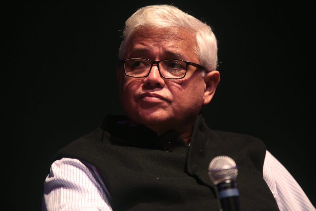 Amitav Ghosh with white hair and glasses, in a dark waistcoat, with a microphone in front of him