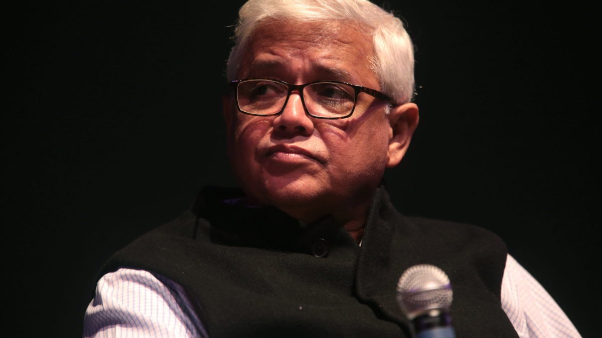 Amitav Ghosh with white hair and glasses, in a dark waistcoat, with a microphone in front of him