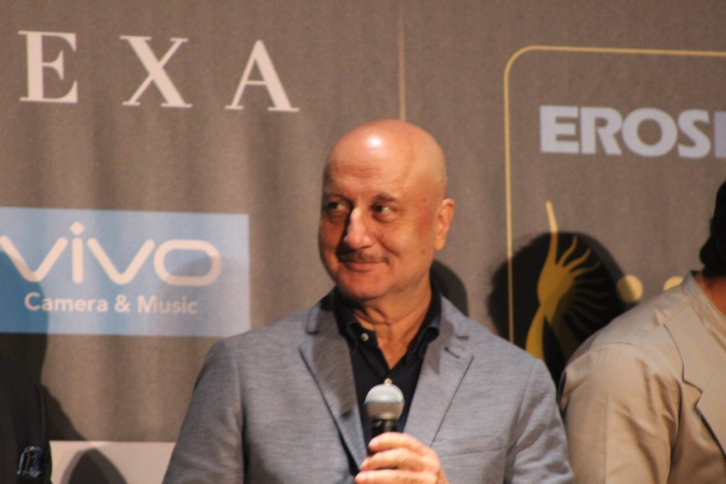 Anupam Kher smiling in a grey jacket, holding a microphone at an IIFA press conference