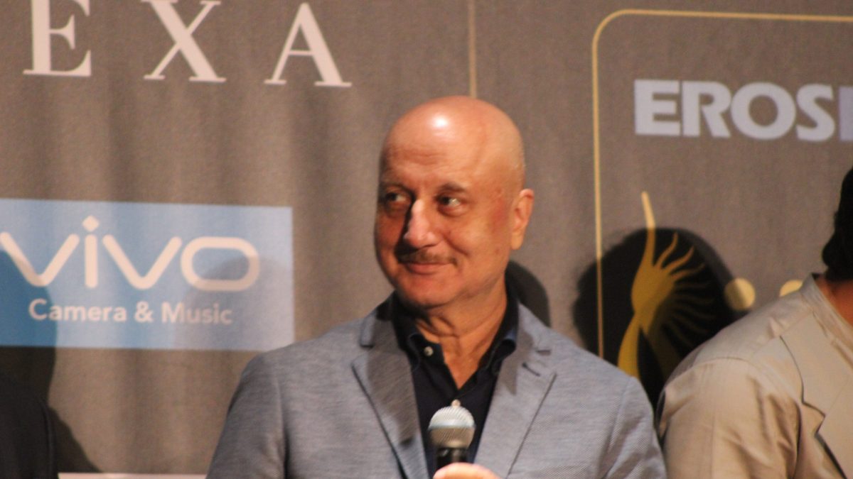 Anupam Kher smiling in a grey jacket, holding a microphone at an IIFA press conference