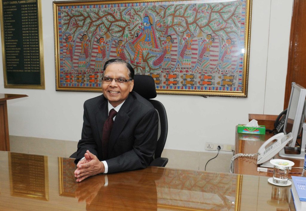 Arvind Panagariya in a dark suit seated at his desk, smiling, with a Madhubani painting behind him