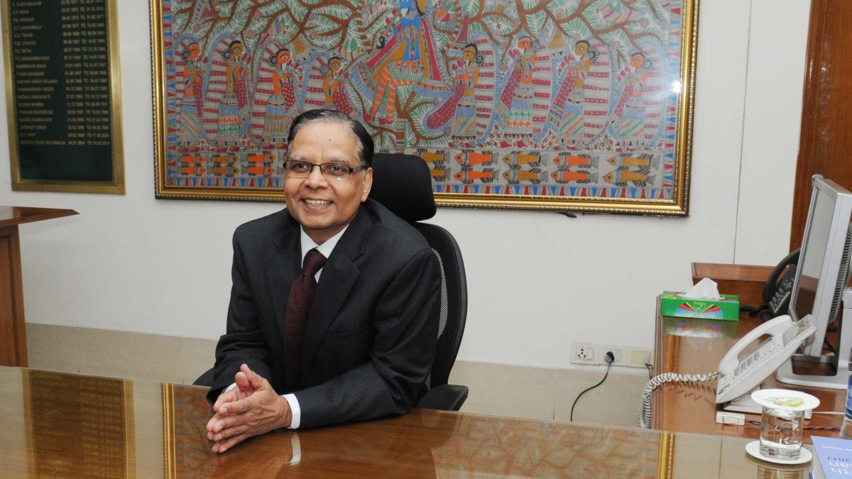 Arvind Panagariya in a dark suit seated at his desk, smiling, with a Madhubani painting behind him