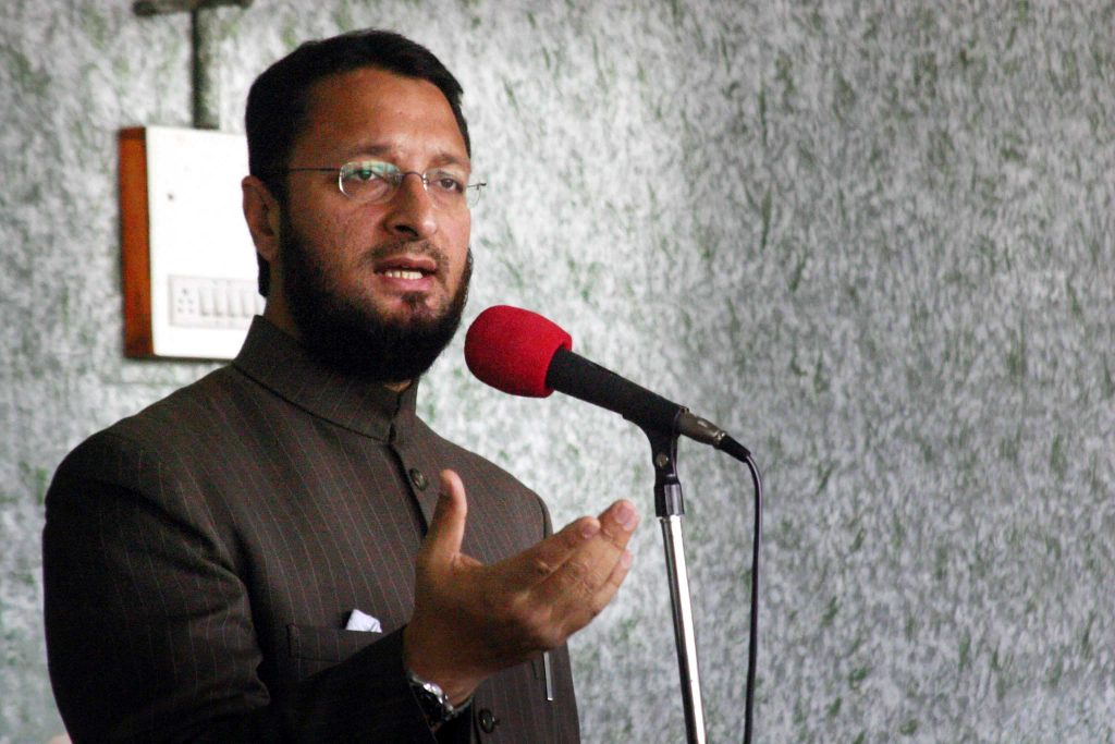 Asaduddin Owaisi with a beard, wearing a cap and sherwani, speaking at microphones