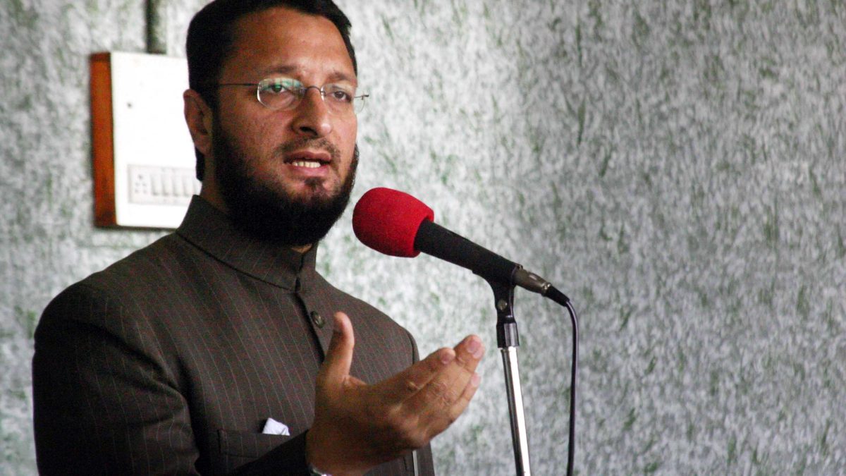 Asaduddin Owaisi with a beard, wearing a cap and sherwani, speaking at microphones