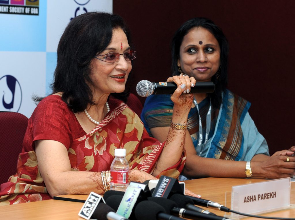 Asha Parekh in a red sari speaking into a microphone at a press conference