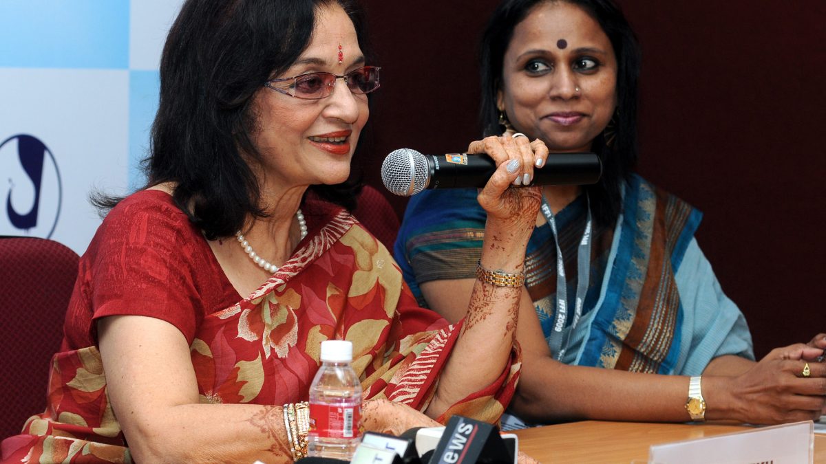 Asha Parekh in a red sari speaking into a microphone at a press conference