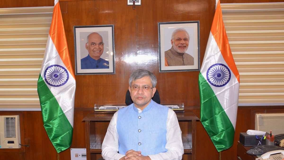 Ashwini Vaishnaw in a blue kurta seated at a desk between two Indian flags