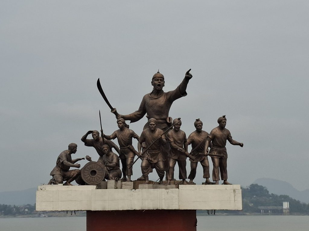 A bronze statue of Lachit Borphukan pointing forward with his soldiers and a cannon, on a platform beside the Brahmaputra