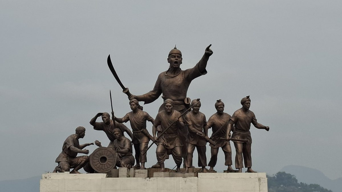 A bronze statue of Lachit Borphukan pointing forward with his soldiers and a cannon, on a platform beside the Brahmaputra