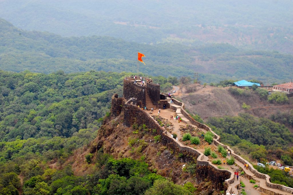 The stone ramparts of Pratapgad fort along a ridge above forested hills, with a saffron flag flying