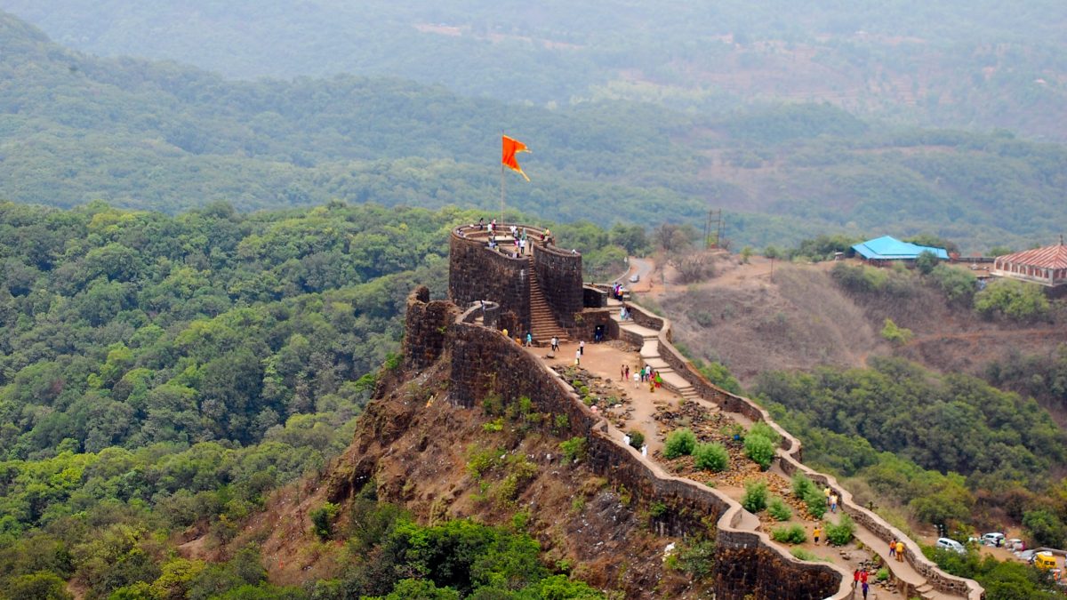 The stone ramparts of Pratapgad fort along a ridge above forested hills, with a saffron flag flying