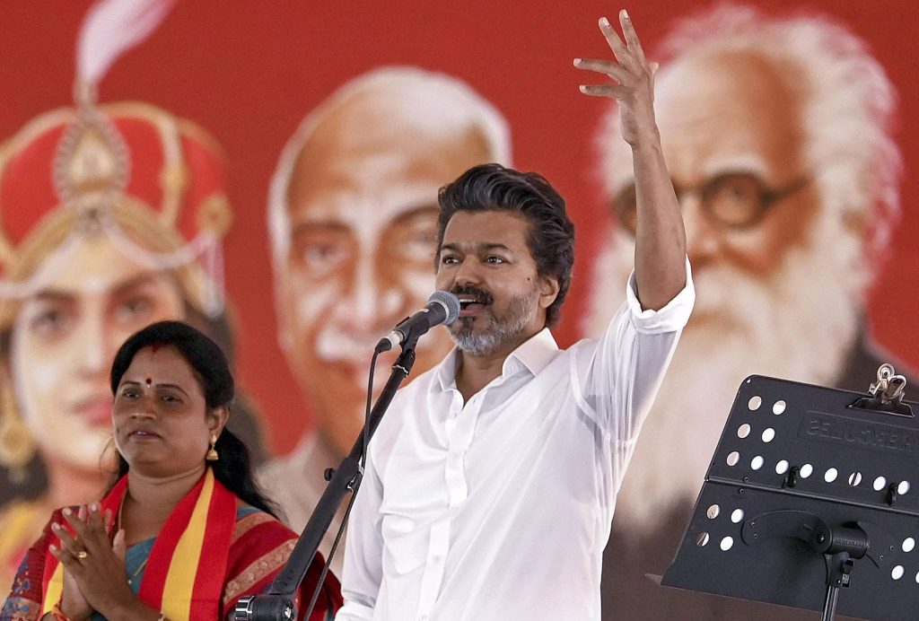 A speaker raising his arm at a microphone in front of a red party backdrop, with a woman applauding beside him