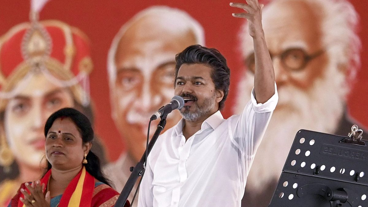 A speaker raising his arm at a microphone in front of a red party backdrop, with a woman applauding beside him