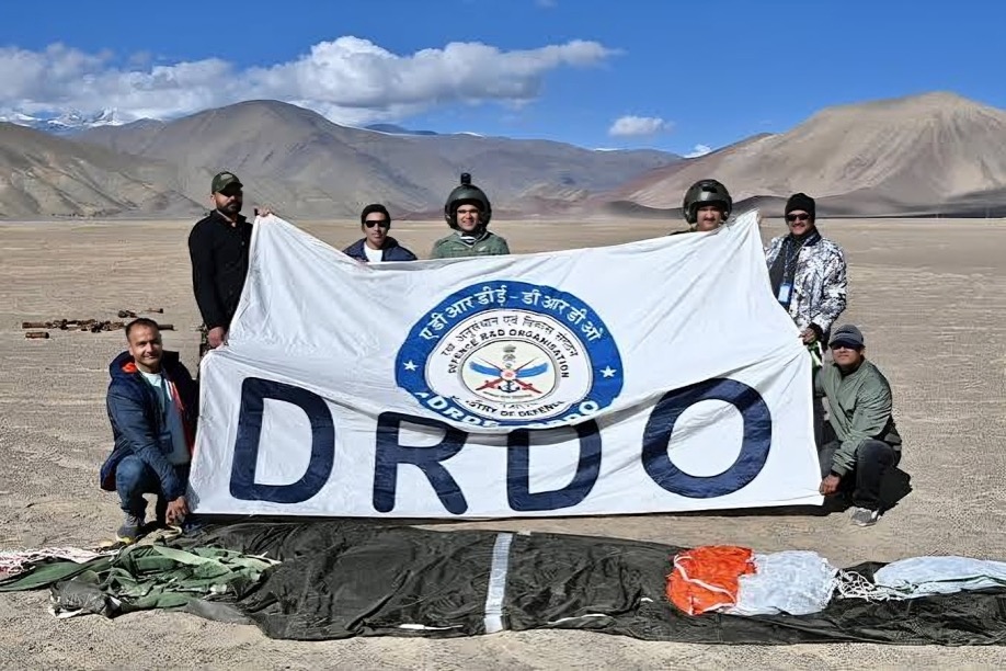 Scientists holding a DRDO banner on a high-altitude plain with mountains behind and a parachute on the ground