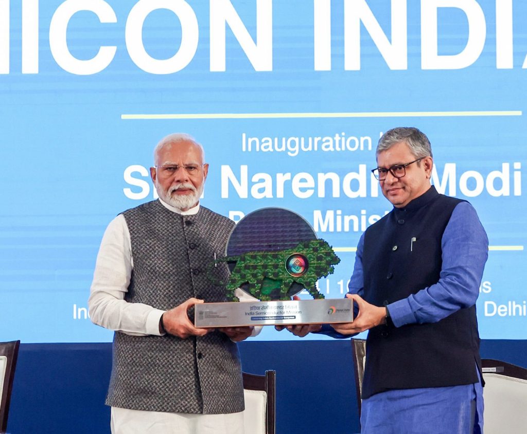 Two men holding an India Semiconductor Mission memento in front of a Semicon India banner