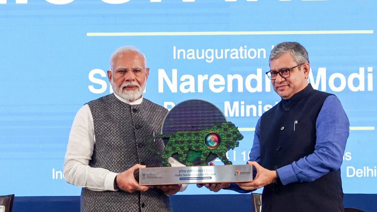 Two men holding an India Semiconductor Mission memento in front of a Semicon India banner