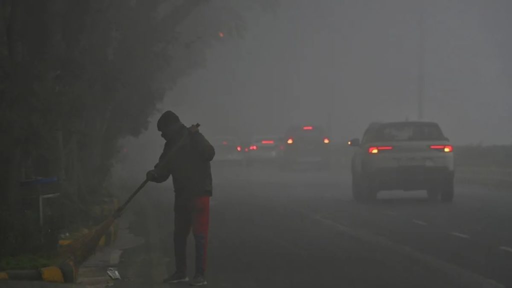 A person sweeping a road in thick smog with a car and tail lights in the background