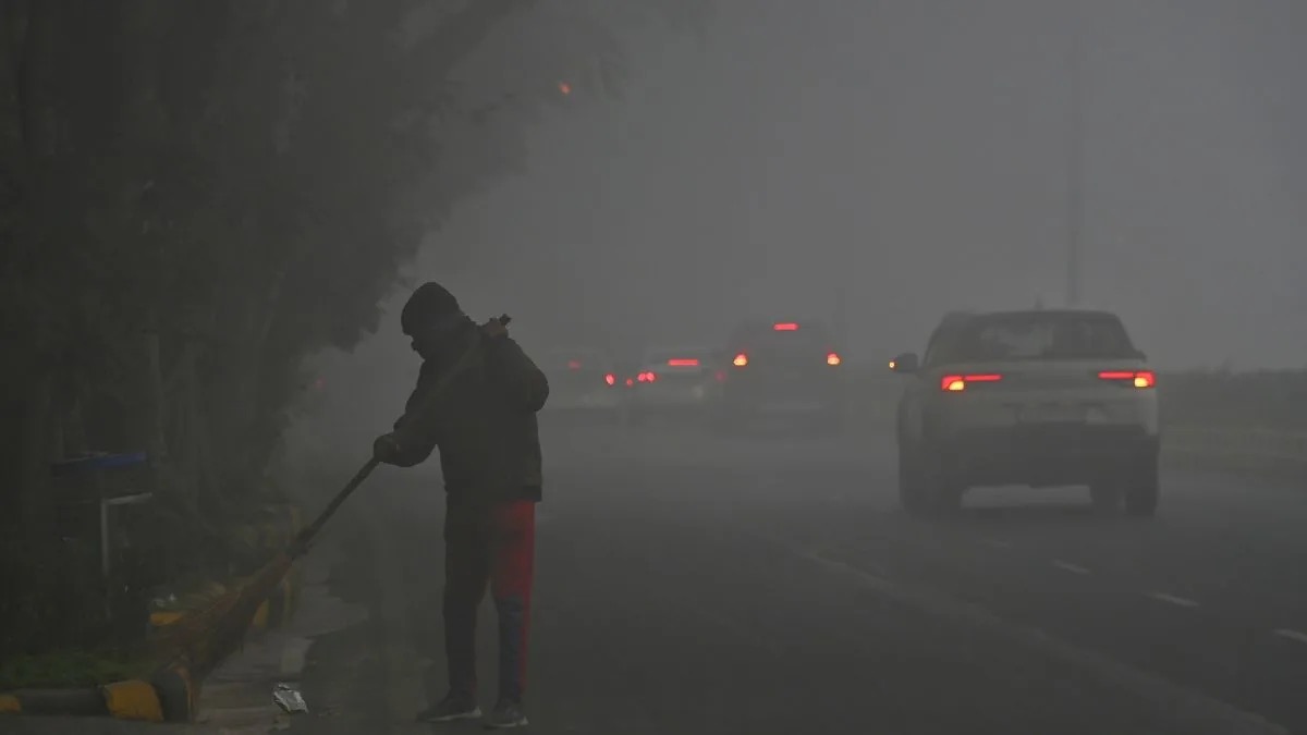 A person sweeping a road in thick smog with a car and tail lights in the background