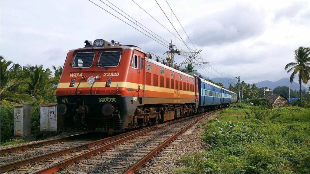 An Indian Railways electric locomotive pulling blue passenger coaches through green countryside