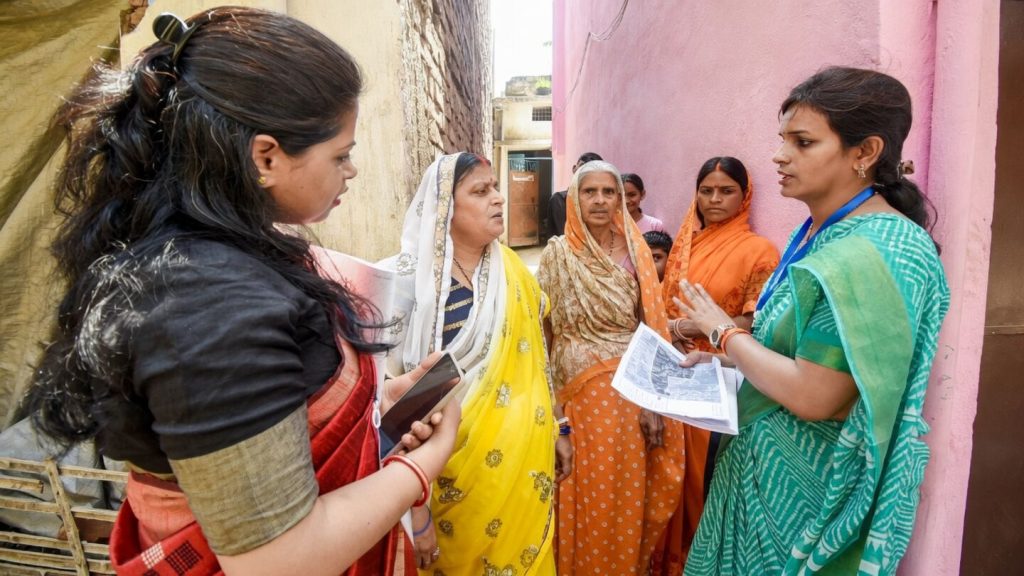 A census enumerator with a phone and papers speaking to a group of women in a lane