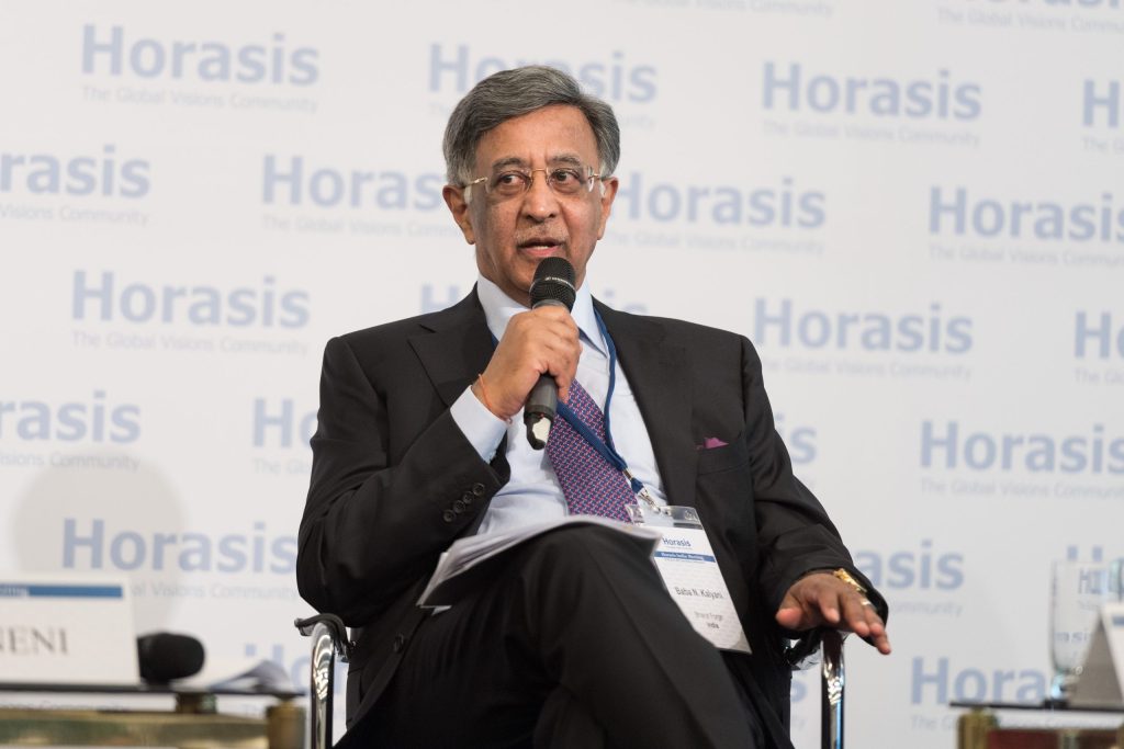 Baba Kalyani in glasses and a dark suit seated on stage holding a microphone