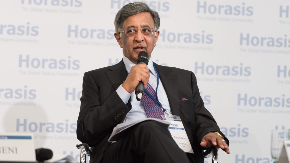 Baba Kalyani in glasses and a dark suit seated on stage holding a microphone
