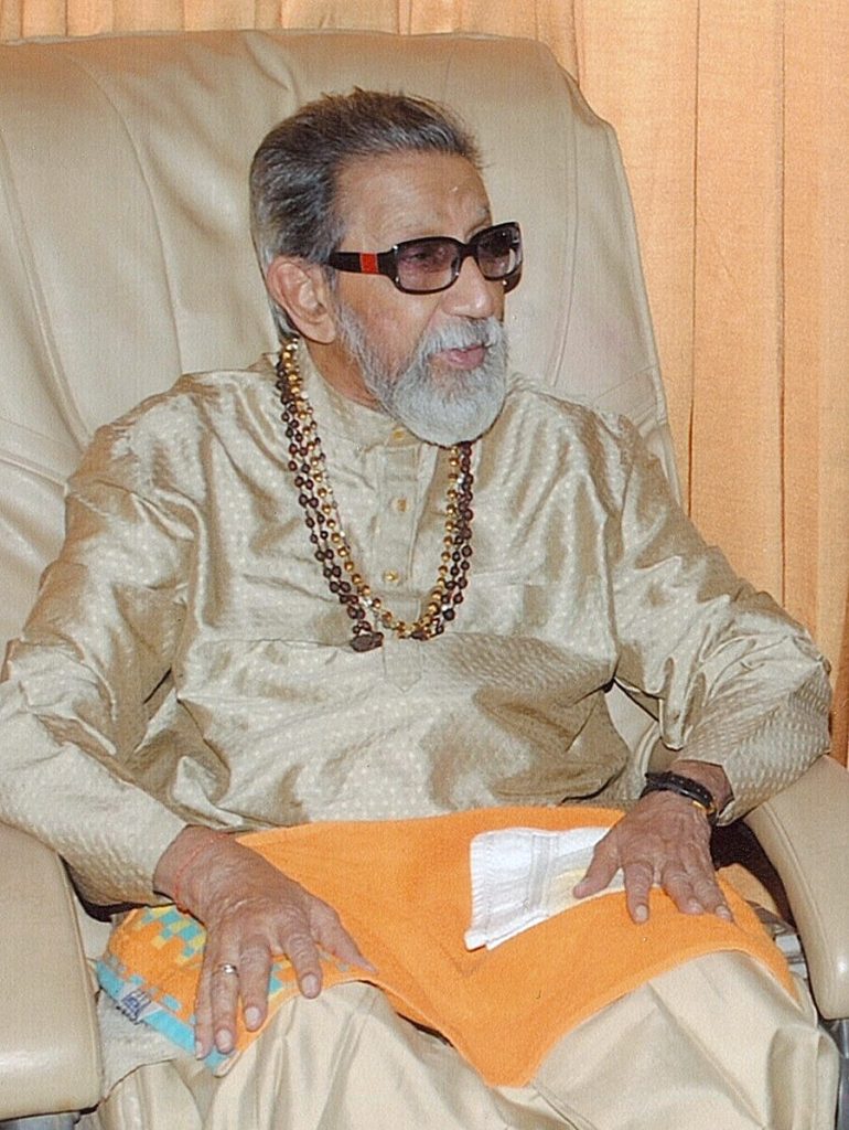 An elderly Bal Thackeray with a white beard and dark glasses, seated in a cream silk kurta with prayer beads