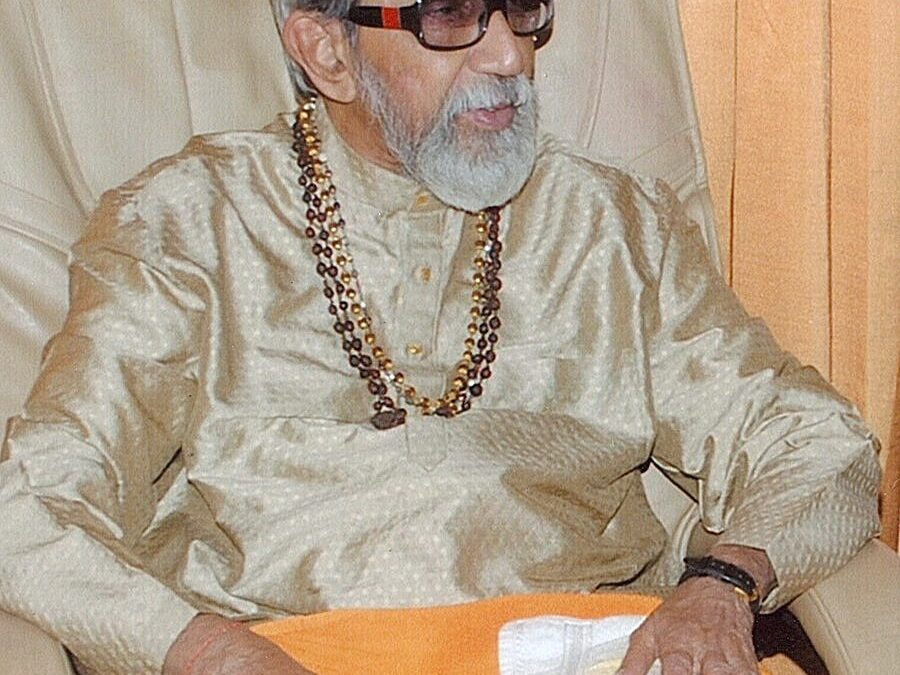 An elderly Bal Thackeray with a white beard and dark glasses, seated in a cream silk kurta with prayer beads