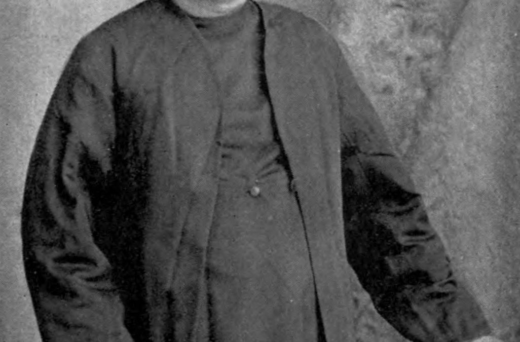 Old photograph of Bankim Chandra Chatterjee standing in a long coat and turban, holding a book