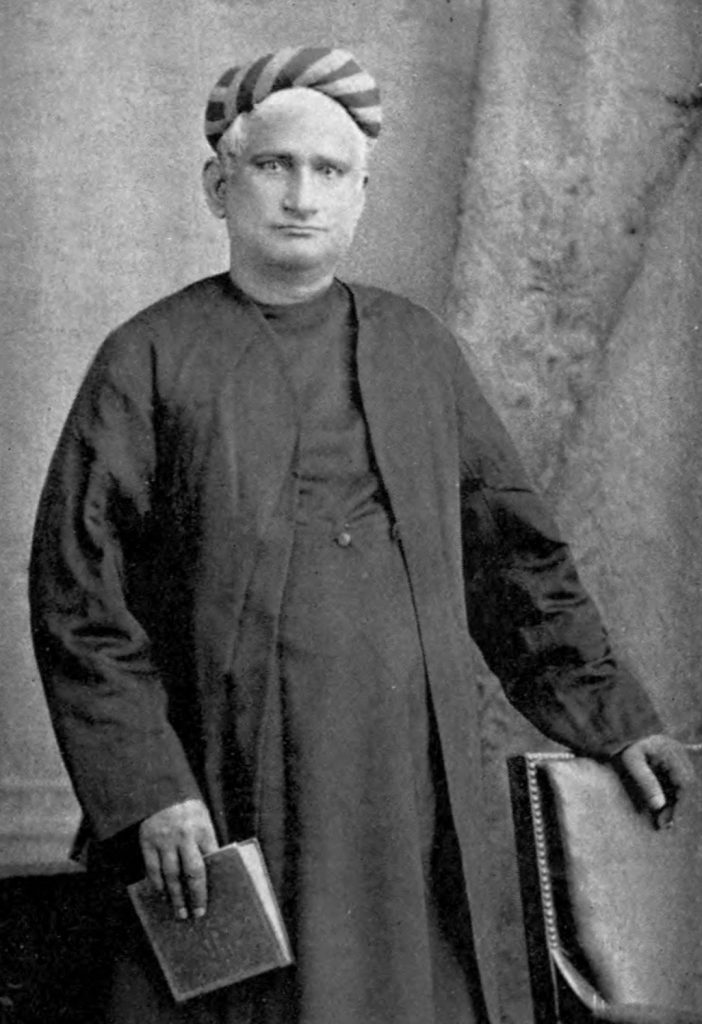 Old photograph of Bankim Chandra Chatterjee standing in a long coat and turban, holding a book